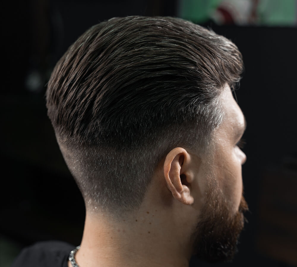 Rear side view of a professional mid-fade haircut with the hair on top slicked back, showcasing clean blending and a groomed beard.